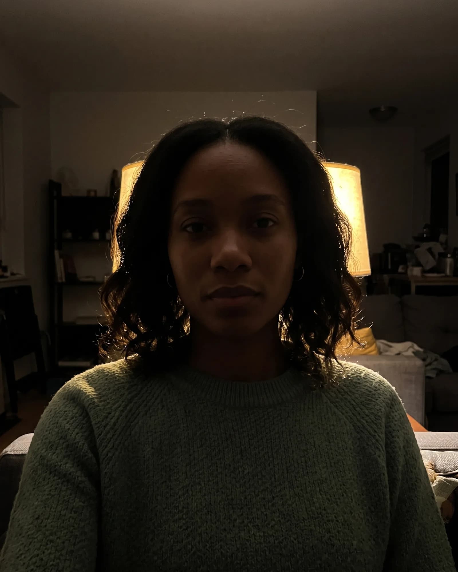 The same woman sitting in a dark room, a lamp glowing behind her head and her face mostly unlit.
